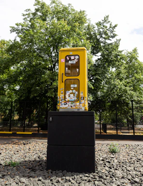 A yellow phone booth is mounted on a black pedestal in an outdoor setting. The booth is covered with various colorful stickers and is surrounded by a gravel surface. Tall green trees are visible in the background, and a fence can be seen lining the perimeter.