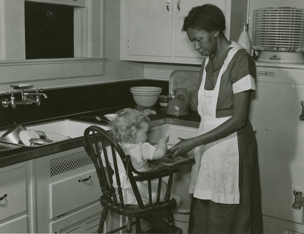 grayscale photography of woman standing in front of baby in highchair