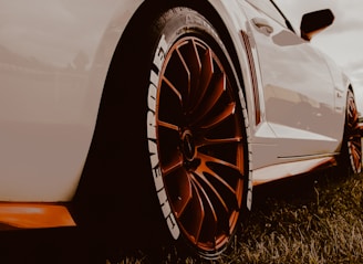 Close-up view of a car's side, showcasing its large, stylish wheel with an intricate rim design. The tire features bold white lettering against a backdrop of a sleek, white car body. The setting appears to be outdoors as the car is parked on grass.