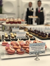 An elegant dessert table featuring an assortment of beautifully crafted pastries and macarons. In the foreground, raspberry, prosecco, and rose petal macarons are arranged neatly with delicate decorations. In the background, various other desserts are displayed on stands. Two servers, wearing formal attire, are visible in the backdrop.