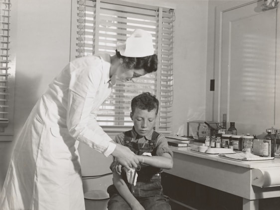 A nurse in a white uniform is tending to a young boy's injured arm in a medical setting. The room contains medical supplies and instruments on a counter in the background, suggesting a traditional clinic or hospital environment. Both figures are focused on the task, creating a scene of care and attention.