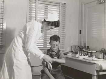 A nurse in a white uniform is tending to a young boy's injured arm in a medical setting. The room contains medical supplies and instruments on a counter in the background, suggesting a traditional clinic or hospital environment. Both figures are focused on the task, creating a scene of care and attention.