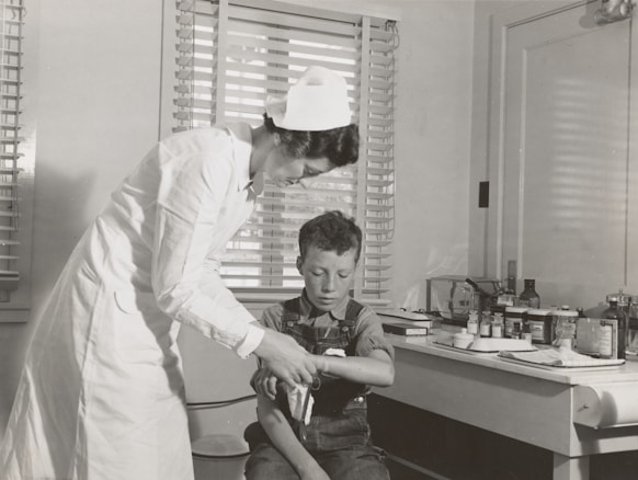 A nurse in a white uniform is tending to a young boy's injured arm in a medical setting. The room contains medical supplies and instruments on a counter in the background, suggesting a traditional clinic or hospital environment. Both figures are focused on the task, creating a scene of care and attention.