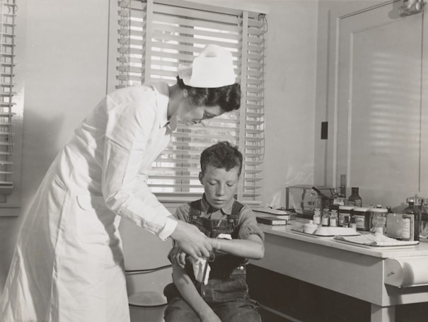 A nurse in a white uniform is tending to a young boy's injured arm in a medical setting. The room contains medical supplies and instruments on a counter in the background, suggesting a traditional clinic or hospital environment. Both figures are focused on the task, creating a scene of care and attention.