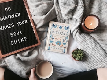 A cozy setting featuring a letter board with the phrase 'Do whatever makes your soul shine' and a book titled 'The Little Book of Hygge'. A lit candle and a small succulent plant add to the warmth. A gray knit blanket is spread out, and a person holds a mug, suggesting a comfortable and relaxed atmosphere.