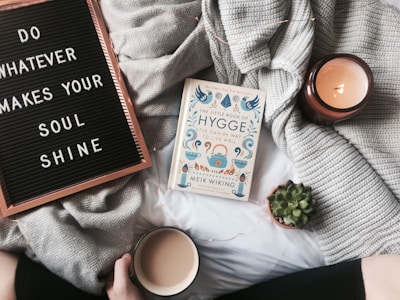 A cozy setting featuring a letter board with the phrase 'Do whatever makes your soul shine' and a book titled 'The Little Book of Hygge'. A lit candle and a small succulent plant add to the warmth. A gray knit blanket is spread out, and a person holds a mug, suggesting a comfortable and relaxed atmosphere.