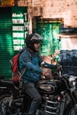 A person wearing a helmet and a blue jacket is riding a black Royal Enfield motorcycle. The individual carries a red and gray backpack. In the background, there is a green sign indicating a Tibetan Handicraft Cooperative Society, and a green door is visible on a brick wall.