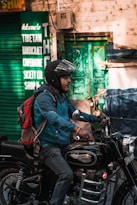 A person wearing a helmet and a blue jacket is riding a black Royal Enfield motorcycle. The individual carries a red and gray backpack. In the background, there is a green sign indicating a Tibetan Handicraft Cooperative Society, and a green door is visible on a brick wall.