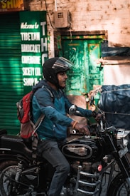 A person wearing a helmet and a blue jacket is riding a black Royal Enfield motorcycle. The individual carries a red and gray backpack. In the background, there is a green sign indicating a Tibetan Handicraft Cooperative Society, and a green door is visible on a brick wall.