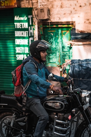 A person wearing a helmet and a blue jacket is riding a black Royal Enfield motorcycle. The individual carries a red and gray backpack. In the background, there is a green sign indicating a Tibetan Handicraft Cooperative Society, and a green door is visible on a brick wall.
