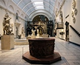 An elegant museum gallery featuring classical sculptures, including human figures depicted in dynamic poses. The gallery has high arched ceilings with skylights, and the floor is covered with ornate tiles. The walls are adorned with additional sculptural art, and the space is illuminated by natural light streaming through the roof.