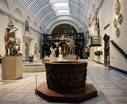 An elegant museum gallery featuring classical sculptures, including human figures depicted in dynamic poses. The gallery has high arched ceilings with skylights, and the floor is covered with ornate tiles. The walls are adorned with additional sculptural art, and the space is illuminated by natural light streaming through the roof.