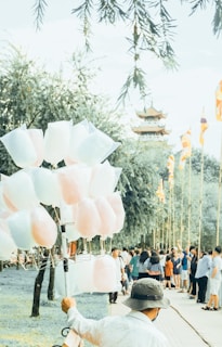 A vendor carries sticks of colorful cotton candy in a public park, surrounded by people walking and socializing. Tall trees with green foliage line the path, and flags are visible in the distance, possibly indicating a cultural or festive event. A pagoda-style structure can be seen in the background.