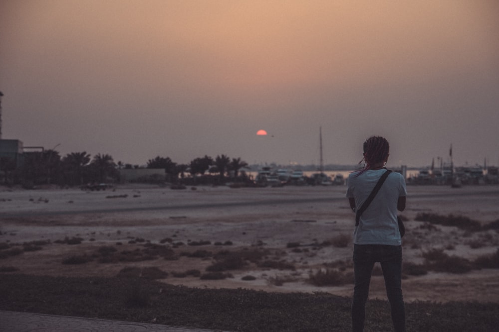 Homme portant un t-shirt blanc et un jean bleu debout près du champ au coucher du soleil