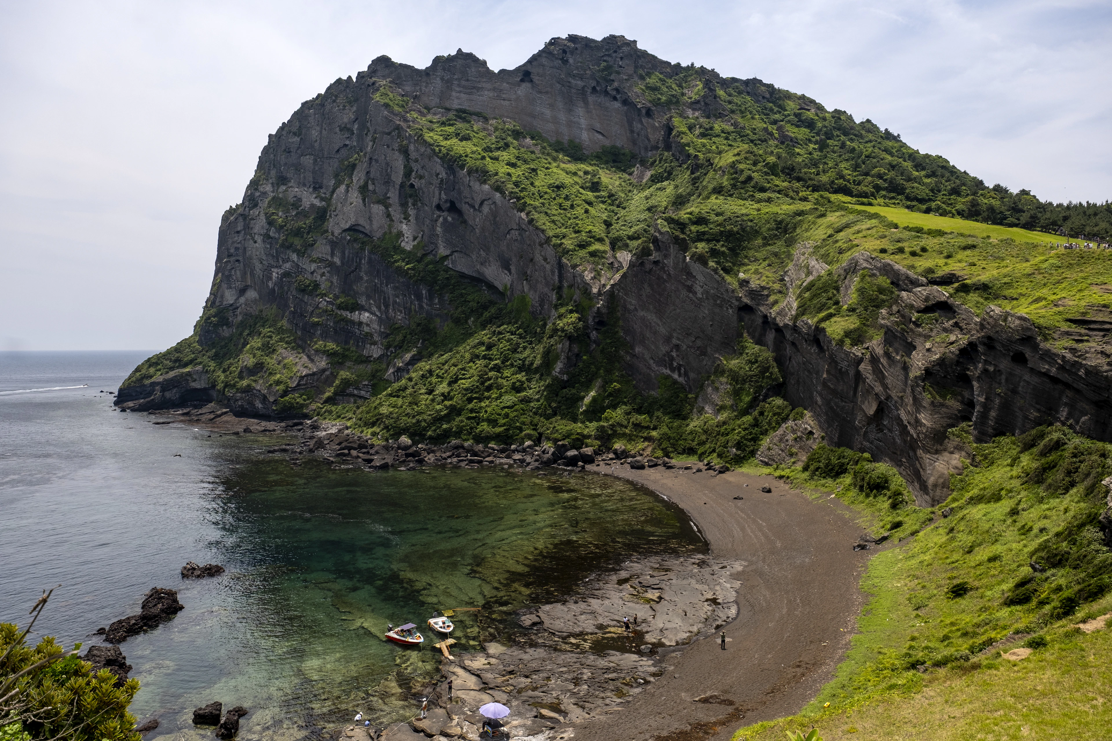 The green volcanic cliffs of Seongsan Ilchulbong on Jeju Island curving around a black-pebble cove and clear turquoise water