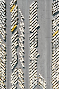 An aerial view of numerous buses parked in a large lot, arranged diagonally in rows. The buses predominantly feature white and yellow colors.