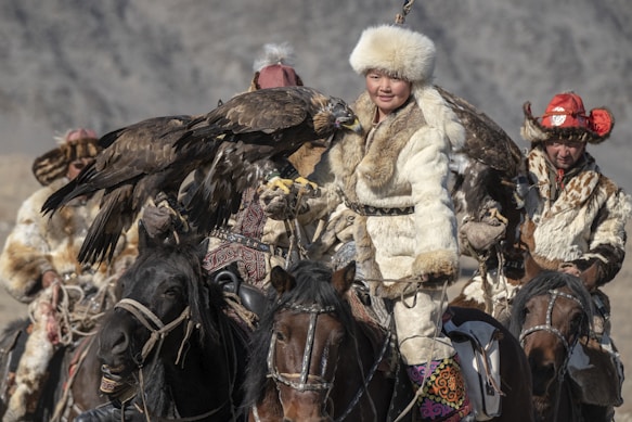 A group of people wearing traditional fur clothing is riding horses. Each person is holding a large bird, possibly an eagle, on their arm. The landscape appears rugged and mountainous, suggesting a remote location. The attire and activity suggest cultural or traditional practices related to eagle hunting.