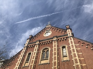 A historic brick church facade with ornate windows and a central clock beneath a cross. The sky above is clear with a few wispy clouds and a visible contrail.