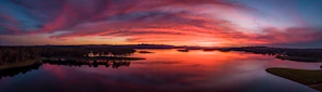 A panoramic view of a sunset over a calm lake, reflecting the fiery sky and distant mountains.