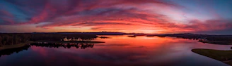 A panoramic view of a sunset over a calm lake, reflecting the fiery sky and distant mountains.