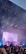 A lively concert scene with bright stage lights and a large screen displaying abstract visuals. The stage has performers and is surrounded by a crowd of enthusiastic fans with raised hands. The sky appears to be twilight, enhancing the colorful lighting.