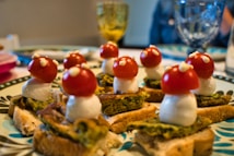A plate features creatively arranged appetizers resembling mushrooms, made from cherry tomatoes, mozzarella balls, and bread with a green spread. The presentation is playful, with small white dots on the tomatoes to mimic mushroom spots. Glasses and a blurred figure are visible in the background, suggesting a dining setup.