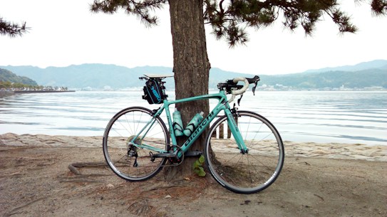 A road bicycle is leaning against a tree by a calm lakeside. The scene includes a background of distant hills and a clear sky, reflected in the still water. The ground is a mixture of sand and rocks.