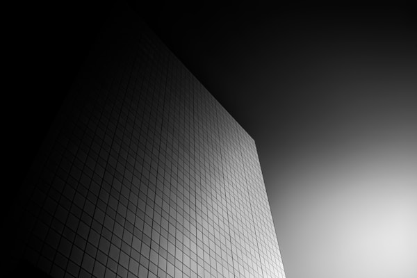 A minimalist, black-and-white photograph of a building's facade. The structure is composed of a grid of square windows, creating a sense of symmetry and order. The lighting adds depth and dimension, with a gradient from dark to light across the image, emphasizing the geometric pattern.