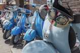 A row of colorful scooters is parked on the street alongside a brick building. The scooters are primarily blue and light gray, with shiny chrome accents. A helmet with an Italian flag design hangs on one of the handlebars. The scene suggests a casual urban setting with a focus on style and retro design.