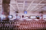 A spacious airport terminal with rows of empty luggage carts lined up in front. Large display signs and digital screens are visible in the background, mounted on the walls. The area is well-lit, showcasing modern architectural elements like high ceilings with grid patterns and metallic pillars.