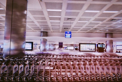 A spacious airport terminal with rows of empty luggage carts lined up in front. Large display signs and digital screens are visible in the background, mounted on the walls. The area is well-lit, showcasing modern architectural elements like high ceilings with grid patterns and metallic pillars.