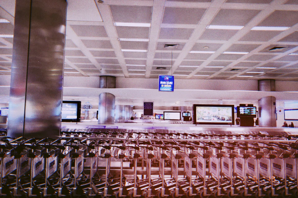 The image depicts a bustling airport terminal with rows of silver luggage carts neatly arranged on the floor, ready for passengers to load their belongings onto them. The terminal is spacious and well-lit, with large windows allowing natural light to flood in and illuminate the scene. A blue sign hangs from the ceiling, possibly providing information or instructions to travelers. In the background, a few people can be seen walking around, likely waiting for their flights or just arriving at the airport after a journey.
