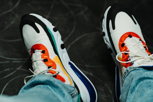 A pair of colorful sports shoes worn with blue jeans. The shoes have a vibrant design, featuring red, orange, white, black, and blue elements. The background appears to be a dark grey surface with subtle, abstract patterns.