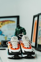 A pair of vibrant Nike sneakers with a mix of orange, white, black, and blue designs are placed on a flat surface. A figurine resembling a superhero is positioned behind the shoes. Framed comic book art is partially visible in the background, adding to a playful and colorful theme.