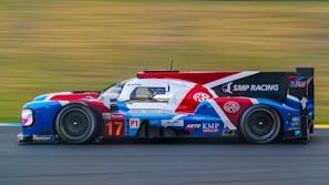 A sleek racing car with a colorful livery, featuring red, white, blue, and black hues, speeds along a blurred racetrack. The vehicle is equipped with sophisticated aerodynamics and is branded with various sponsorship logos. The blurred background conveys a sense of high velocity.