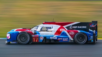 A sleek racing car with a colorful livery, featuring red, white, blue, and black hues, speeds along a blurred racetrack. The vehicle is equipped with sophisticated aerodynamics and is branded with various sponsorship logos. The blurred background conveys a sense of high velocity.