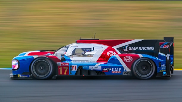 A sleek racing car with a colorful livery, featuring red, white, blue, and black hues, speeds along a blurred racetrack. The vehicle is equipped with sophisticated aerodynamics and is branded with various sponsorship logos. The blurred background conveys a sense of high velocity.