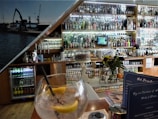 A bar setting with an assortment of alcoholic beverages displayed on shelves behind the counter. A large glass with ice, lemon slices, and a straw is placed on the table in the foreground. A vase with flowers and a cake stand are also visible. Behind the bar, there is a collection of bottles ranging from spirits to mixers. The interior has a cozy ambiance with wooden elements and a photo mural of a dockyard scene.