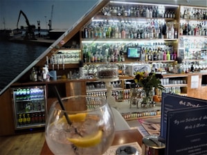 A bar setting with an assortment of alcoholic beverages displayed on shelves behind the counter. A large glass with ice, lemon slices, and a straw is placed on the table in the foreground. A vase with flowers and a cake stand are also visible. Behind the bar, there is a collection of bottles ranging from spirits to mixers. The interior has a cozy ambiance with wooden elements and a photo mural of a dockyard scene.