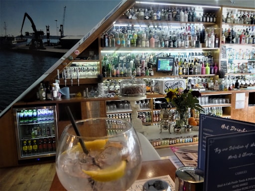 A bar setting with an assortment of alcoholic beverages displayed on shelves behind the counter. A large glass with ice, lemon slices, and a straw is placed on the table in the foreground. A vase with flowers and a cake stand are also visible. Behind the bar, there is a collection of bottles ranging from spirits to mixers. The interior has a cozy ambiance with wooden elements and a photo mural of a dockyard scene.