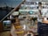 A bar setting with an assortment of alcoholic beverages displayed on shelves behind the counter. A large glass with ice, lemon slices, and a straw is placed on the table in the foreground. A vase with flowers and a cake stand are also visible. Behind the bar, there is a collection of bottles ranging from spirits to mixers. The interior has a cozy ambiance with wooden elements and a photo mural of a dockyard scene.