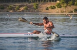 A man is intensely paddling a kayak in a calm body of water. He appears focused, using a double-bladed paddle to propel himself forward. The water splashes around him, and his muscles are visibly strained. Trees line the distant shore, and small floating buoys are scattered in the water under a warm, late afternoon light.
