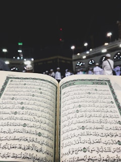 An open Quran is prominently displayed, with Arabic text visible on both pages. The background features an outdoor setting at night, with the Kaaba surrounded by worshippers wearing white clothing, indicative of a religious gathering.