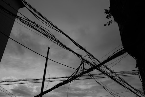 A network of tangled electrical wires crisscrossing against a cloudy sky backdrop. The silhouette of a building corner and some tree branches can be seen. A utility pole stands amid the wires, adding to the urban feel.