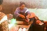 A person is skillfully cutting a fish using a knife on a wooden block. There is a green plastic container on the table and several fish are laid out. The surrounding area appears rustic, with stone walls and plastic bags.