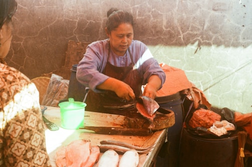 A person is skillfully cutting a fish using a knife on a wooden block. There is a green plastic container on the table and several fish are laid out. The surrounding area appears rustic, with stone walls and plastic bags.
