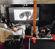 A cozy home theater setup with people lounging on couches and covered in blankets. They are watching a large screen with an animated robot character displayed. The space includes a projector on a table and dim ambient lighting, with a decorative deer head mounted on the wall.