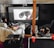 A cozy home theater setup with people lounging on couches and covered in blankets. They are watching a large screen with an animated robot character displayed. The space includes a projector on a table and dim ambient lighting, with a decorative deer head mounted on the wall.