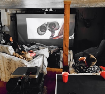 A cozy home theater setup with people lounging on couches and covered in blankets. They are watching a large screen with an animated robot character displayed. The space includes a projector on a table and dim ambient lighting, with a decorative deer head mounted on the wall.