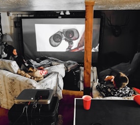A cozy home theater setup with people lounging on couches and covered in blankets. They are watching a large screen with an animated robot character displayed. The space includes a projector on a table and dim ambient lighting, with a decorative deer head mounted on the wall.